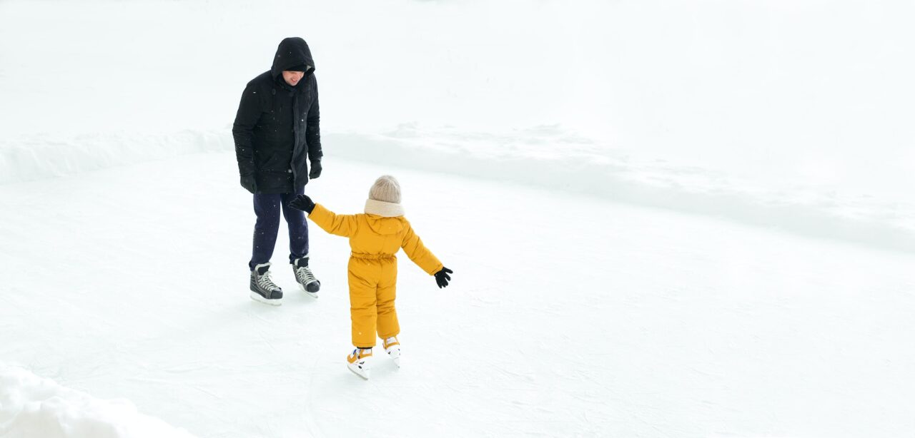 Father and baby are standing on the ice rink opposite each other.